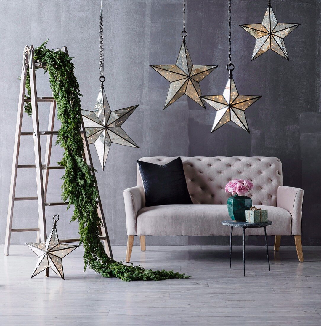 Beleuchtete Deko-Sterne über elegantem Sofa an Metallketten aufgehängt und Zweig-Girlande über Stehleiter drapiert