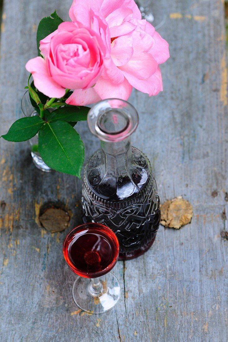 Stielglas neben Karaffe und rosa blühende Rosen in Vase auf Vintage Holzunterlage