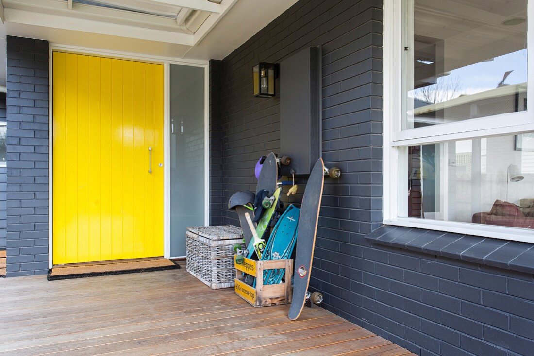 Überdachter Eingangsbereich mit gelber Haustür und grauem Sichtmauerwerk, Holzterrasse und diverse Skateboards in Holzkiste gestapelt