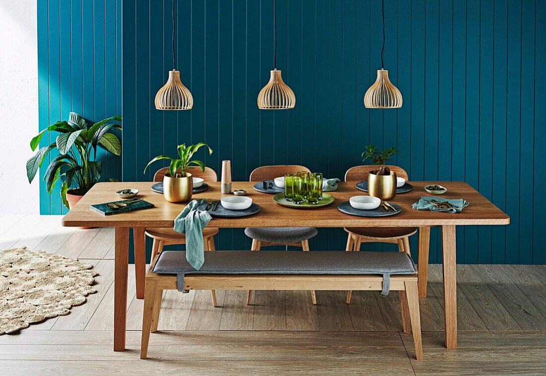 Laid dining table with wooden bench under pendant lights in front of petrol-colored wooden paneling