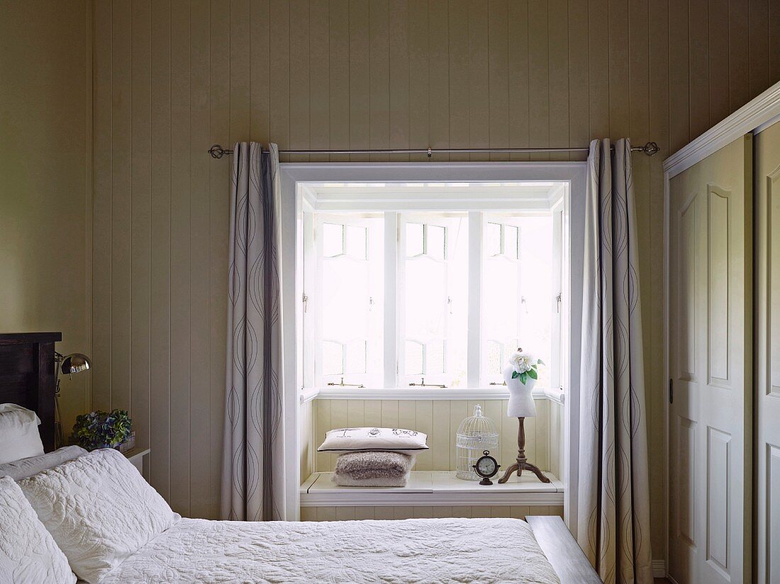Blick über Doppelbett mit weisser Tagesdecke in holzverschaltem Schlafzimmer auf Fensternische