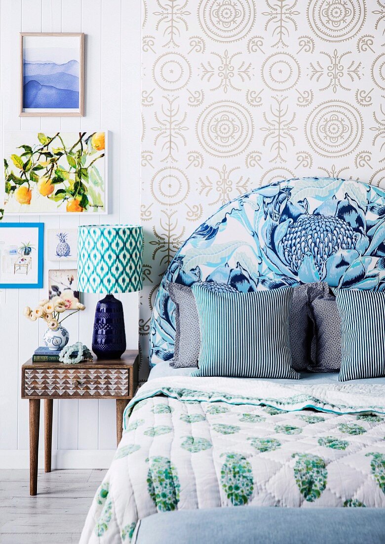 Bedroom in a mix of patterns; Bed with blue upholstered headboard in front of pattern wallpaper and retro bedside table with table lamp