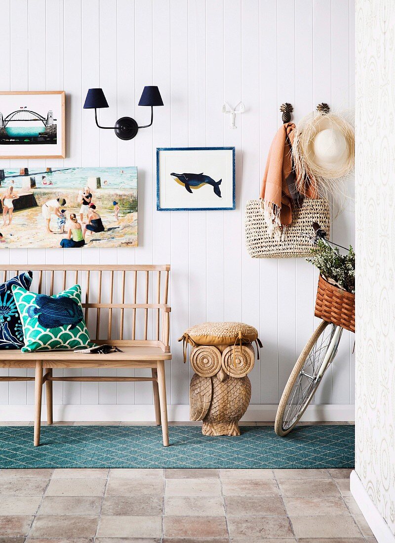 Hausflur mit Holzbank neben geschnitzter Eulenfigur als Hocker vor weisser Wandverkleidung mit Strandbild und maritimem Flair