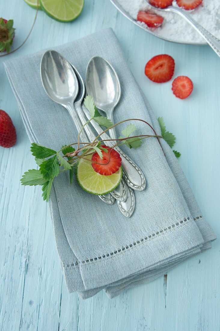 Silberlöffel auf Serviette mit Erdbeerranken, Erdbeeren und Limette