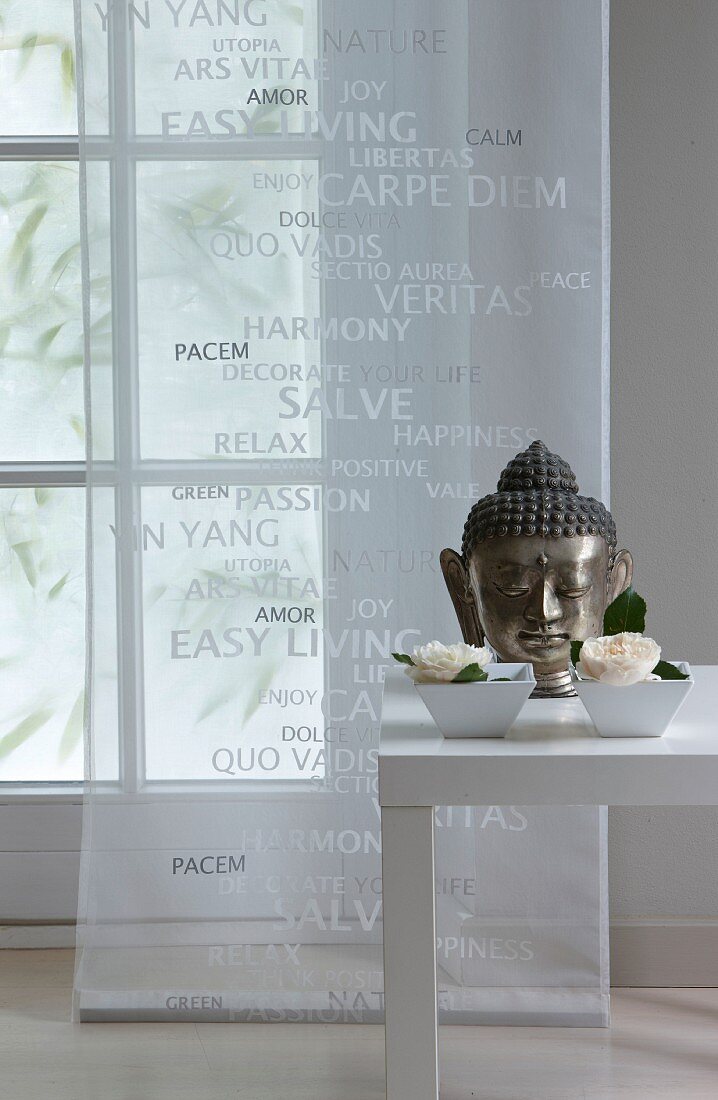 Head of Buddha and flowers in china bowls decorating table in front of window with transparent curtain panel