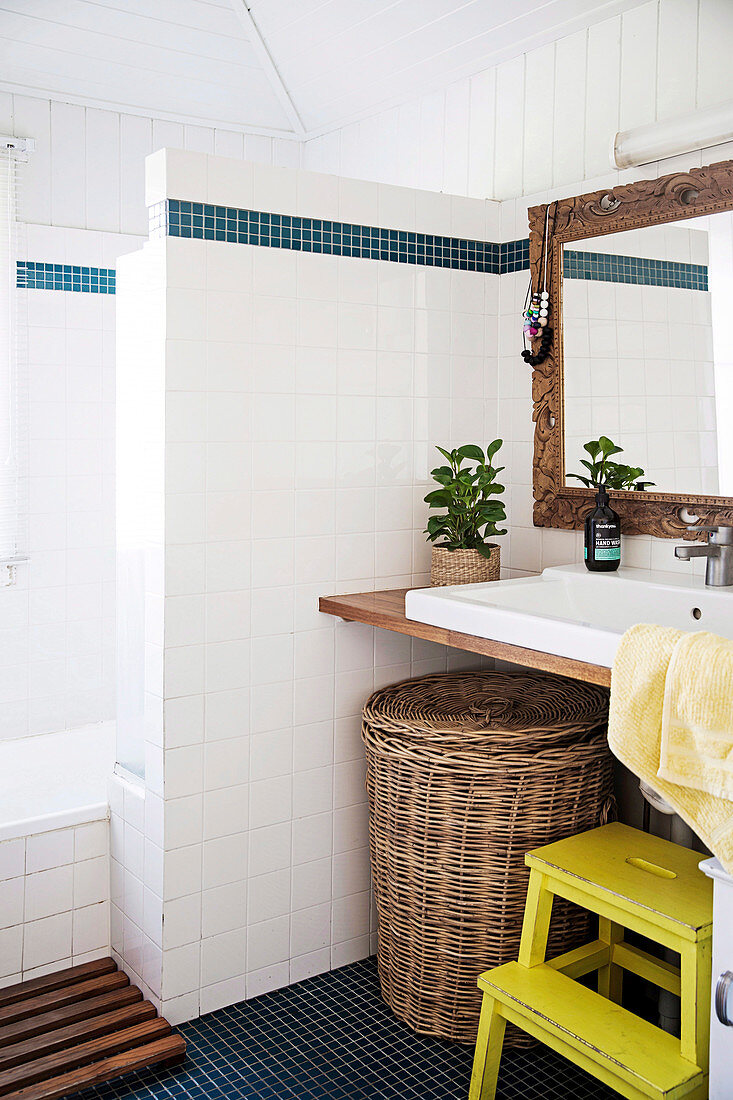 Laundry basket under vanity top next to yellow step stool in white bathroom with Scandinavian flair