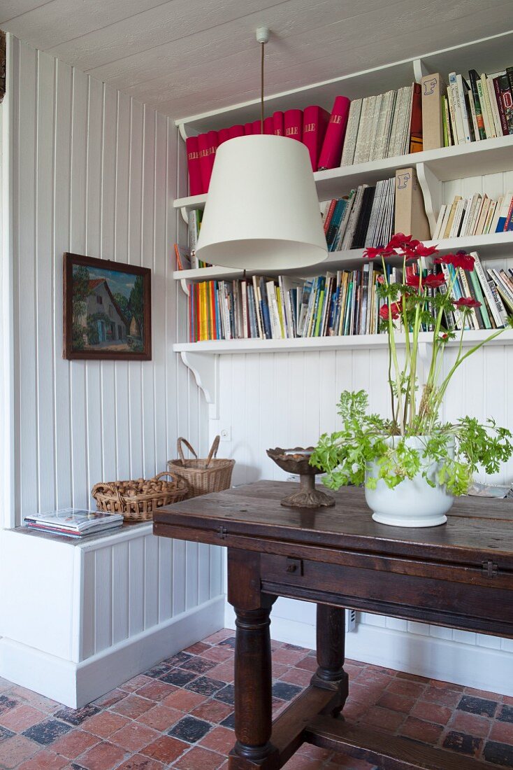 Antiker Konsolentisch mit blühender Topfpflanze vor weisser Holzwand mit Bücherbrettern