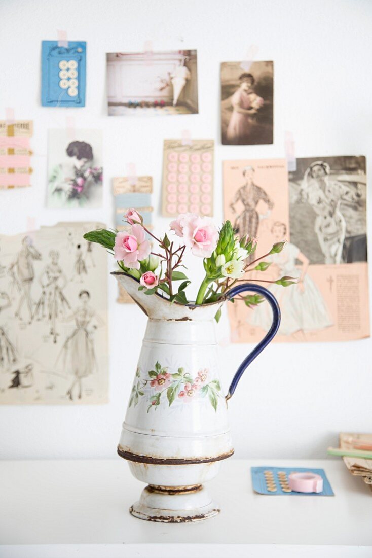 Roses in old enamel jug in front of vintage-style pictures on wall