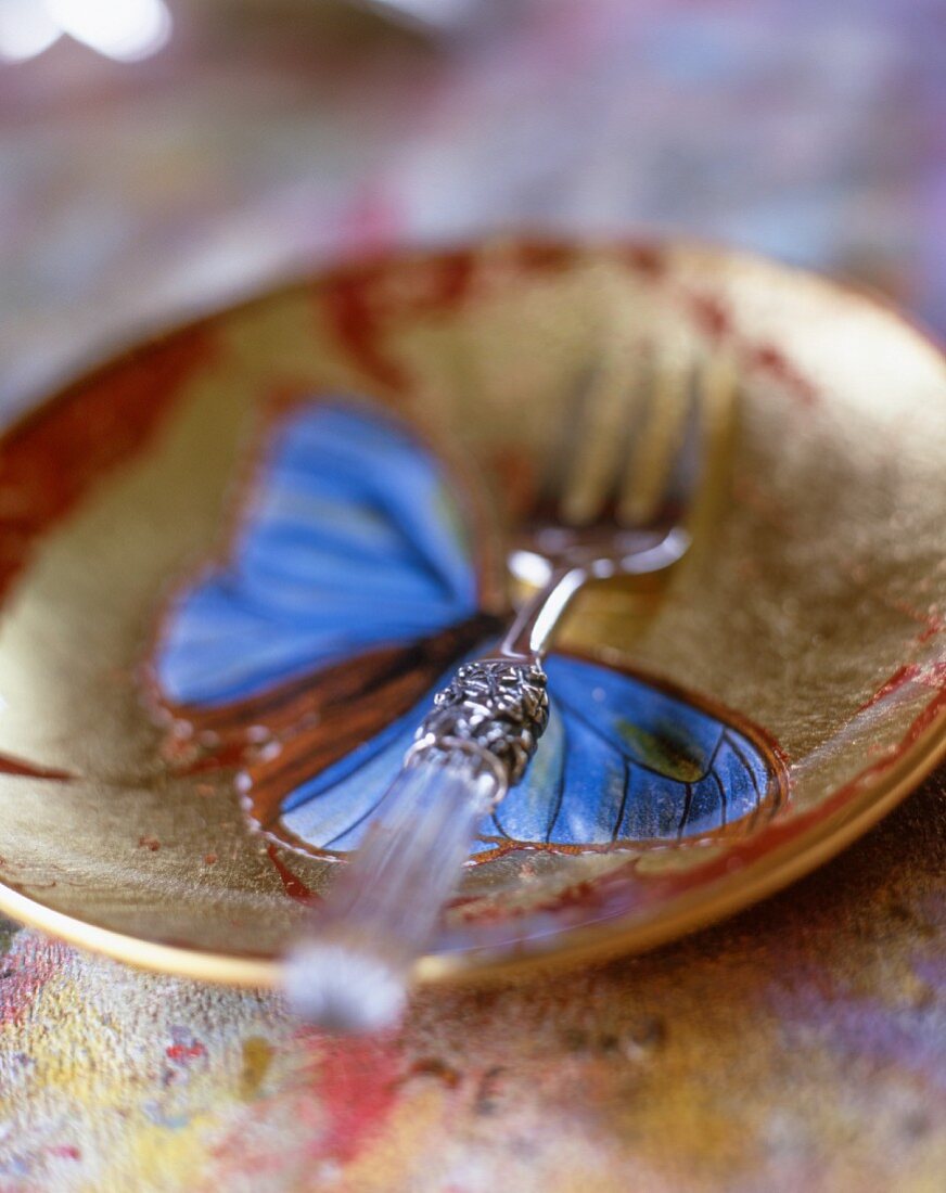 Keramikteller mit Schmetterlingsmotiv, darin eine silberne Gabel mit Glasgriff