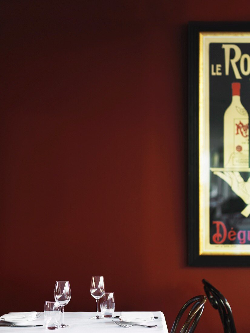 Gedeckter Tisch mit Wein- und Wassergläsern vor dunkelroter Wand mit Retro-Werbeplakat