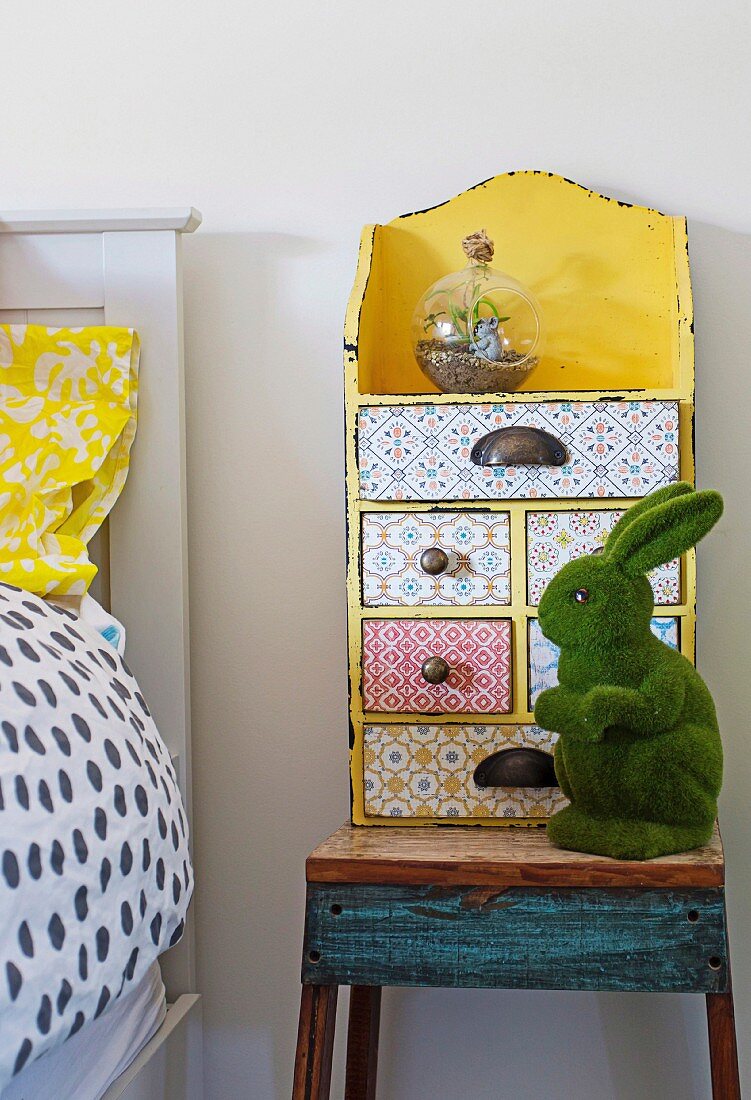 Gelbes Vintage Schubladenkästchen mit verschiedenen Mustern beklebt und grüne Hasenfigur auf rustikalem Nachttisch in Kinderzimmer