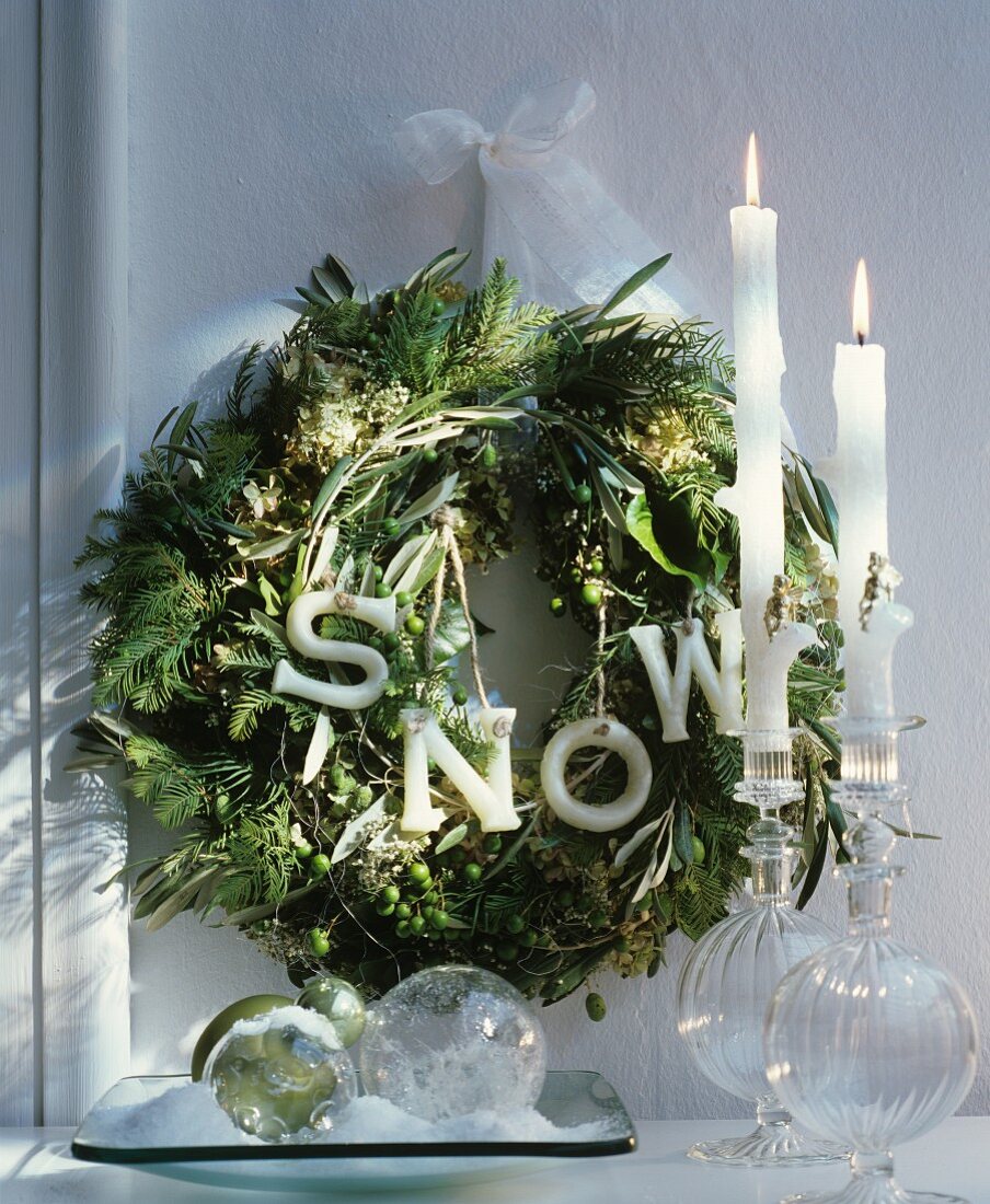 Green wreath with decorative letters and white ribbon behind glass baubles and white candles in elegant glass candlesticks