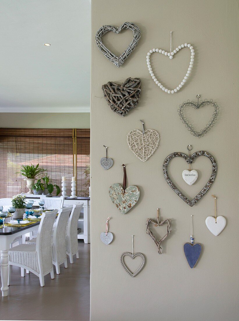 Verschiedene Herzformen aus unterschiedlichen Materialien an grau getönter Wand, seitlich Blick auf gedeckten Esstisch mit weissen Rattan Stühlen