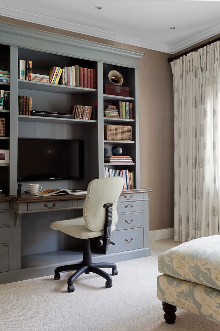 Cremefarbener Bürostuhl vor hellgrauem Wandschrank mit integriertem Schreibtisch und Plasmabildschirm