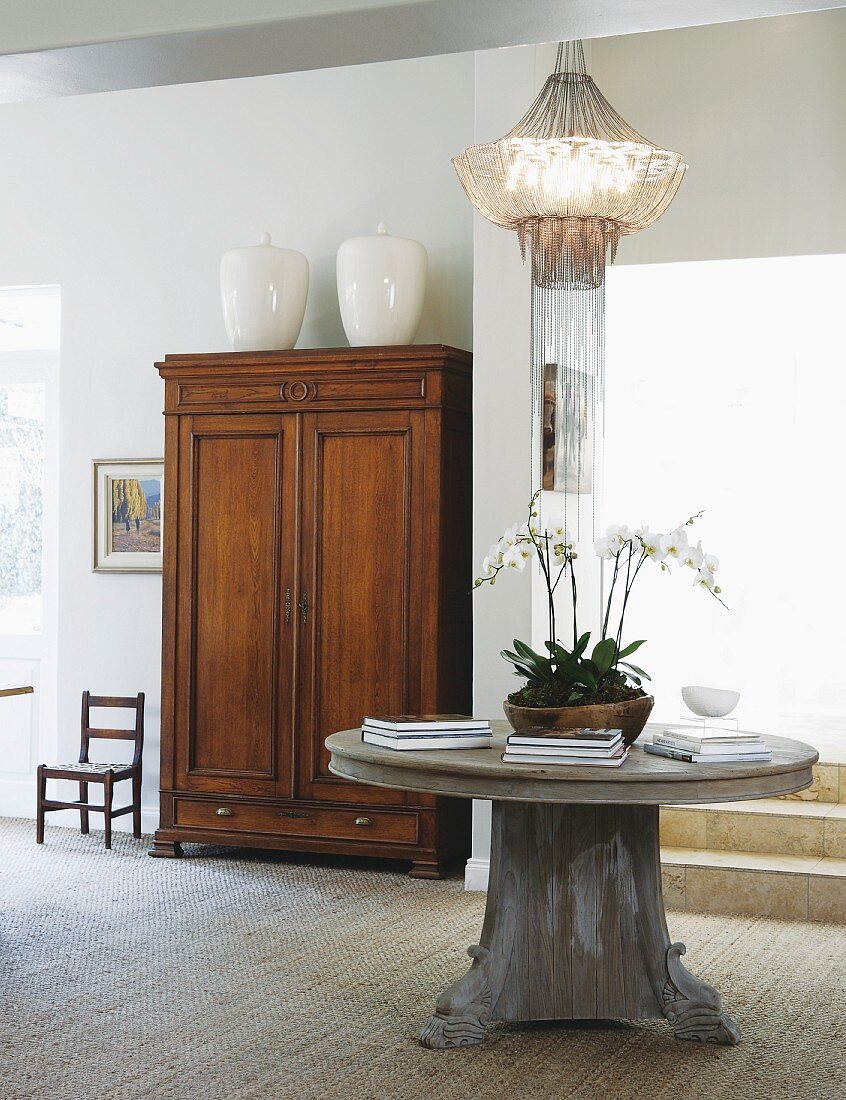 Runder Massivholztisch mit Orchideenschale und Designer-Pendelleuchte in elegantem Wohnraum mit antikem Holzschrank