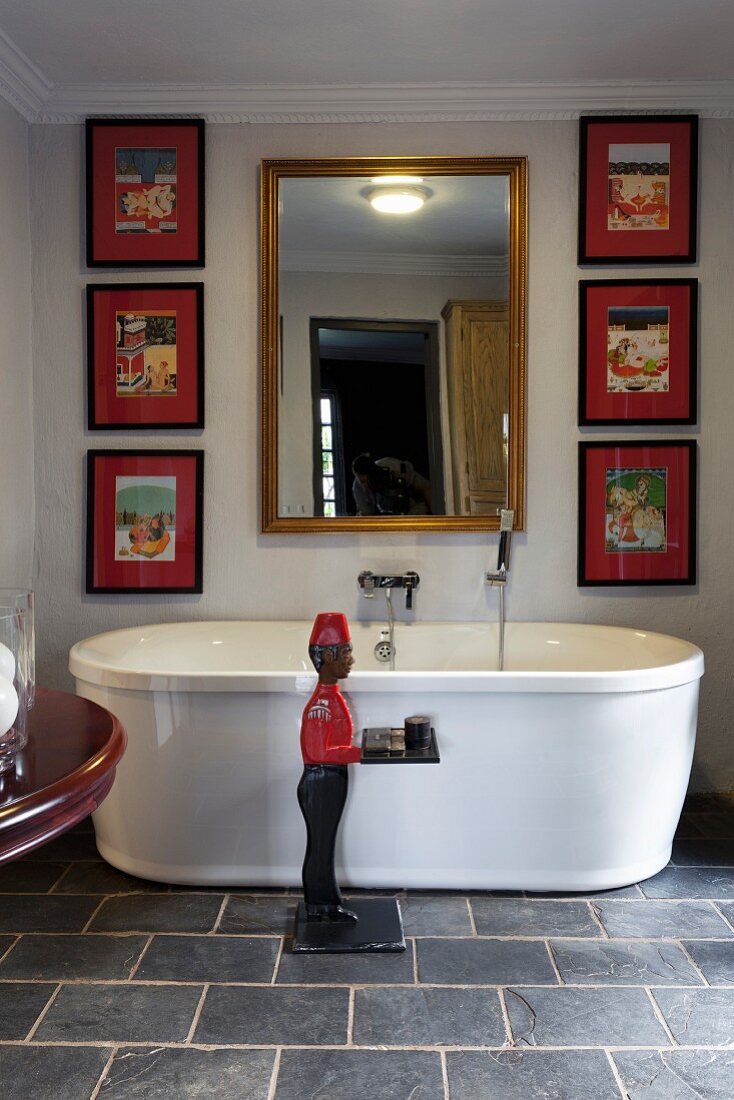 Statue of man next to free-standing bathtub on slate floor below framed mirror and collection of artworks on wall