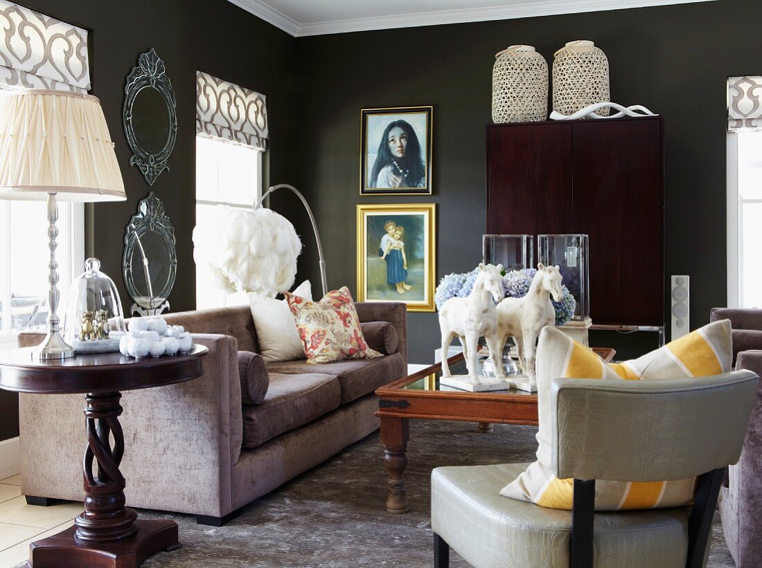 Loungebereich mit Sessel, rundem Beistelltisch aus dunklem Holz und Tischleuchte neben Polstersofa in schwarz getöntem Wohnzimmer