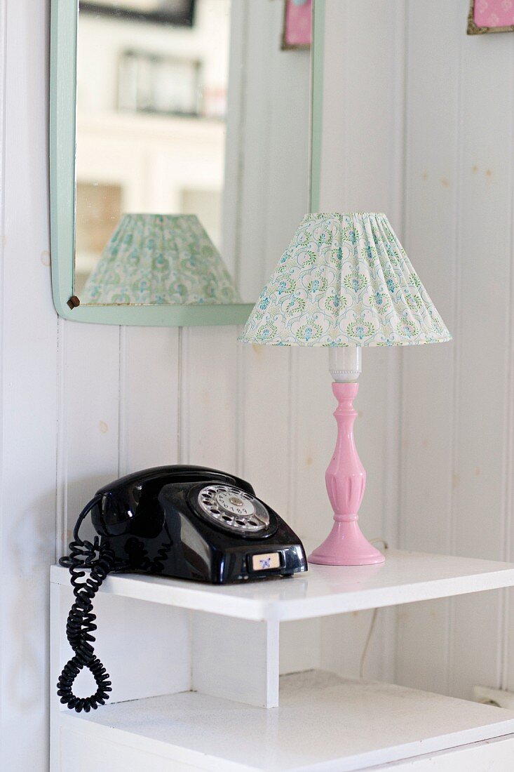 Schwarzes Vintage Telefon und Tischleuchte mit rosa Leuchtenfuß auf weißem Schränkchen vor Wandspiegel