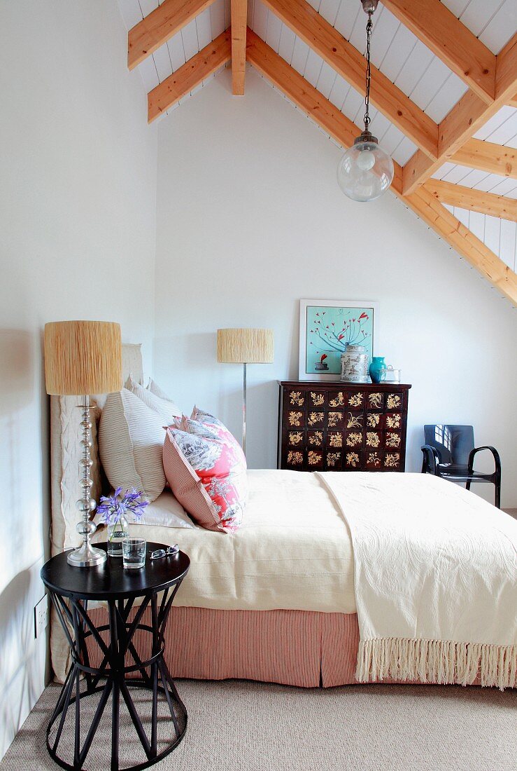 Schwarzer Beistelltisch neben Doppelbett mit heller Tagesdecke im Dachzimmer mit Blick auf Holzbalkendecke
