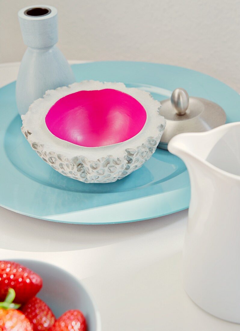 Concrete bowl with magenta inner surface and vase on pale blue porcelain plate, milk jug and bowl of strawberries in foreground