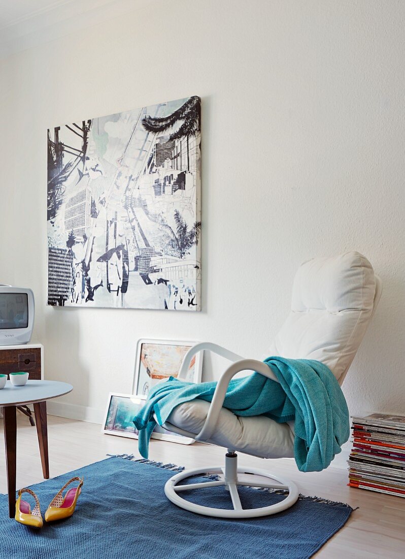 Pale blue blanket on white swivel armchair and modern artwork on wall