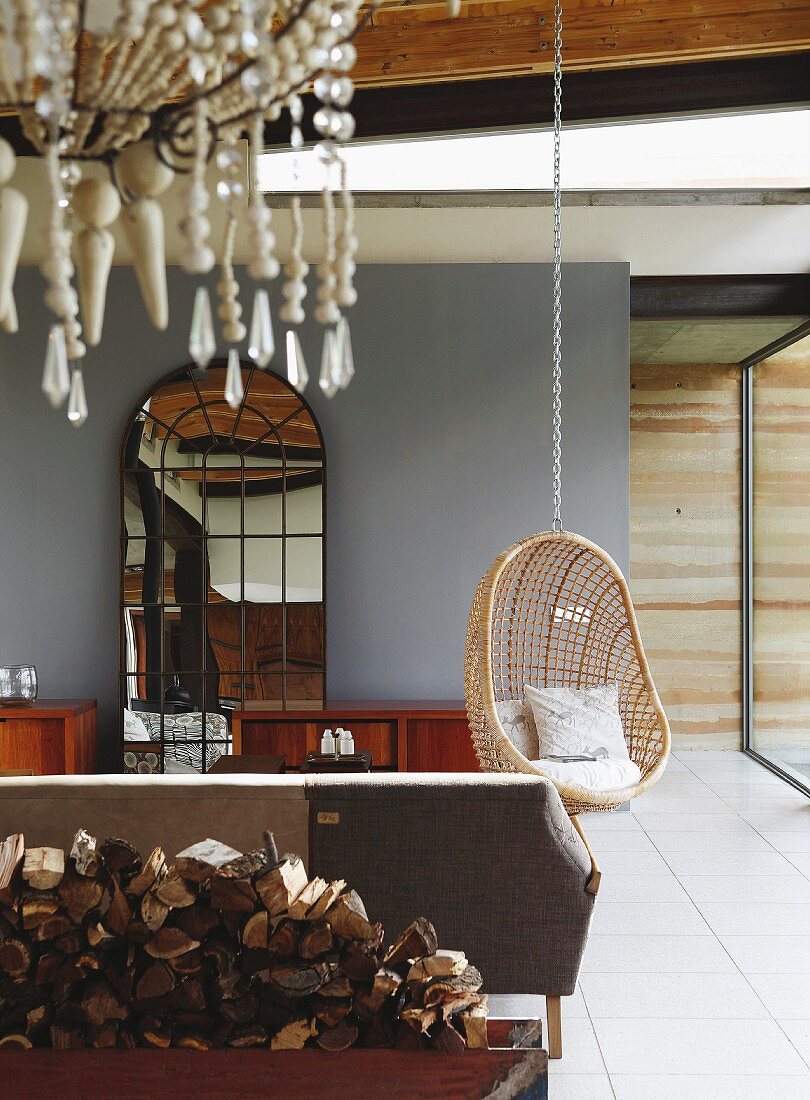 Sofa, Hängekorbstuhl und Standspiegel mit Fenstersprossen in gefliestem Wohnraum; Kronleuchter über Brennholzstapel im Vordergrund