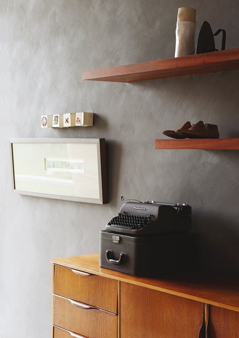 Old typewriter on retro sideboard below ornaments and artworks on wooden floating shelves on grey marbles wall