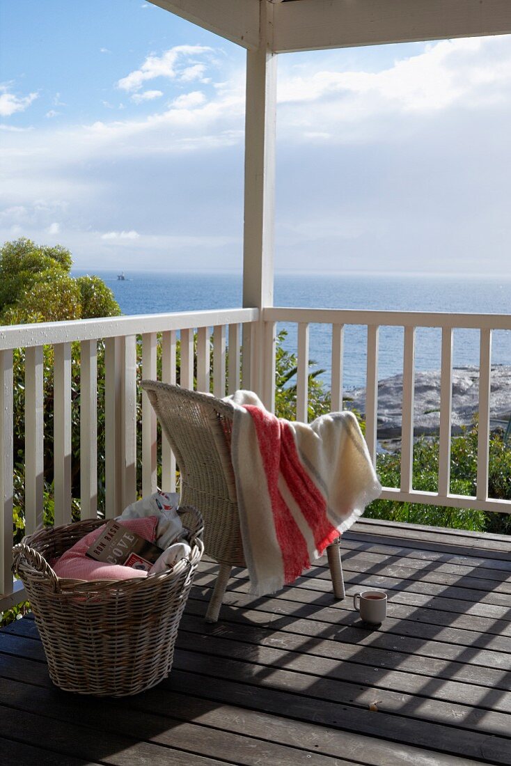 Towel draped over pale wicker chair and basket on sunny wooden deck with sea view