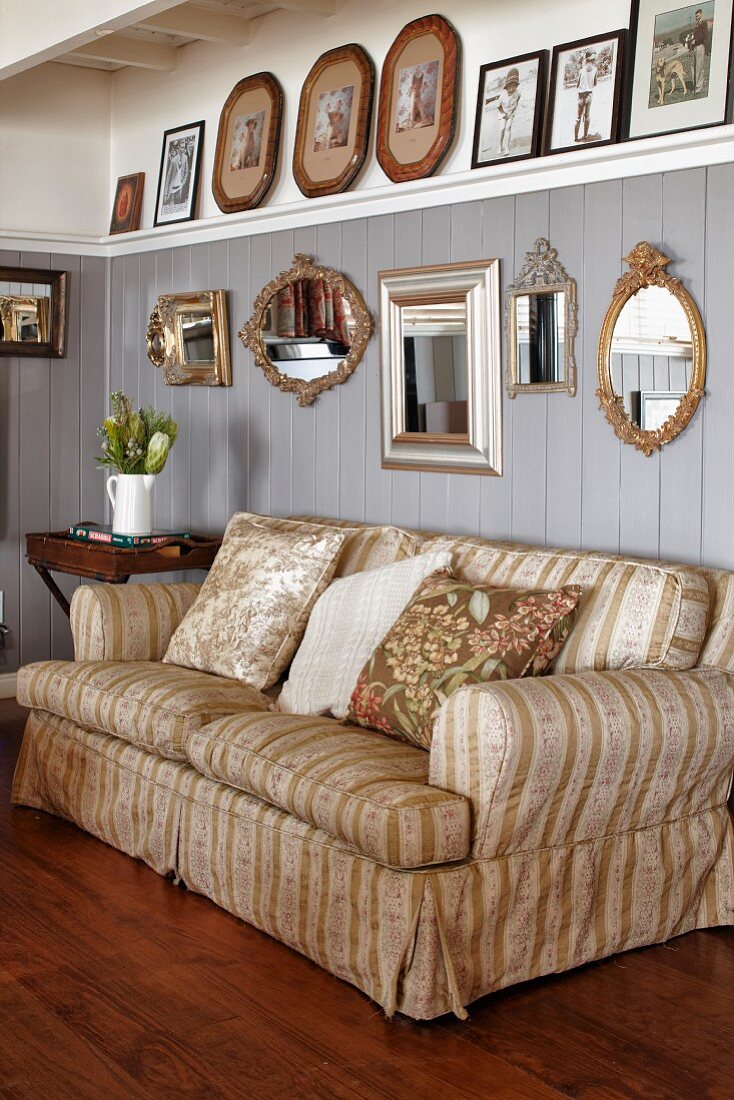 Traditional sofa with pale, striped cover and scatter cushions below framed pictures on grey-painted wooden wall