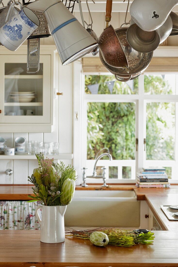 Tropische Pflanzen in weißem Porzellankrug auf Küchentheke, oberhalb aufgehängte Küchenutensilien in ländlicher Küche