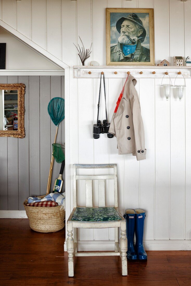 Küchenstuhl vor weisser, holzverkleideter Wand mit offener Garderobe, darüber Portraitbild, im Hauseingang