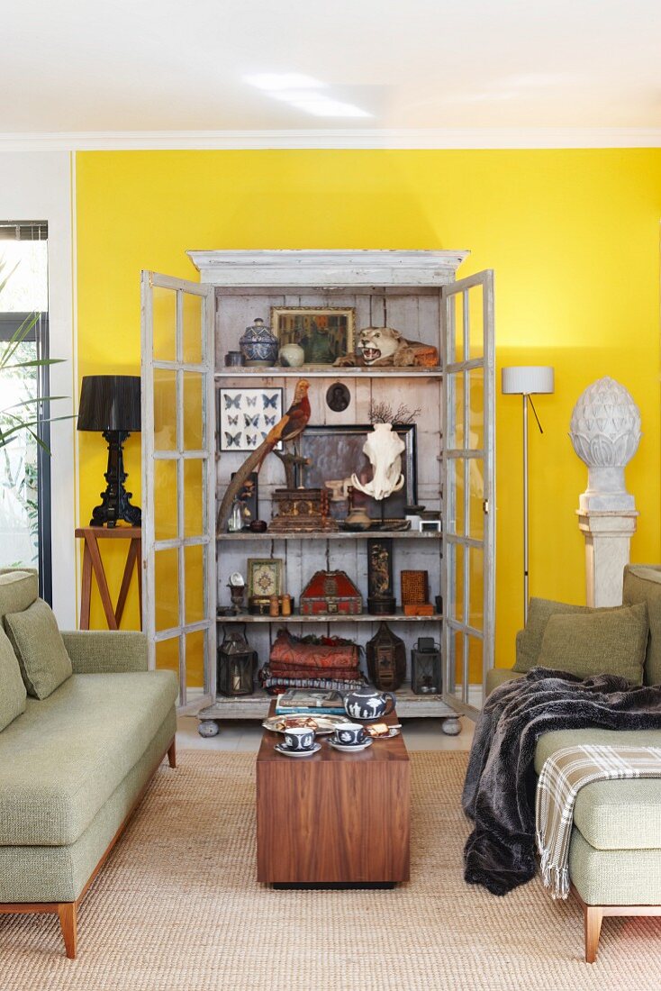 Sofagarnitur mit hellgrünem Bezug im Fiftiesstil und Holztisch vor Vitrinenschrank mit offener Tür, Blick auf antiquarische Sammelstücke im Wohnzimmer mit gelb getönter Wand
