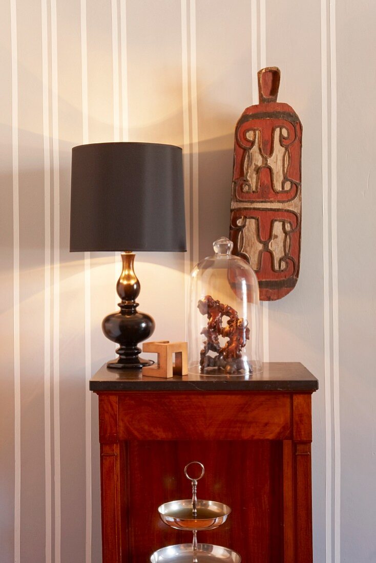Table lamp with black lampshade and glass cover on cabinet below ethnic ornament on wall with white and grey stripes pattern