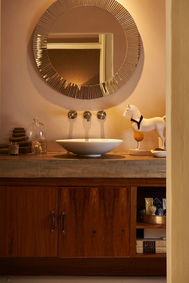 Fitted washstand with basin on concrete counter and wooden base cabinet below round mirror with decorative edge