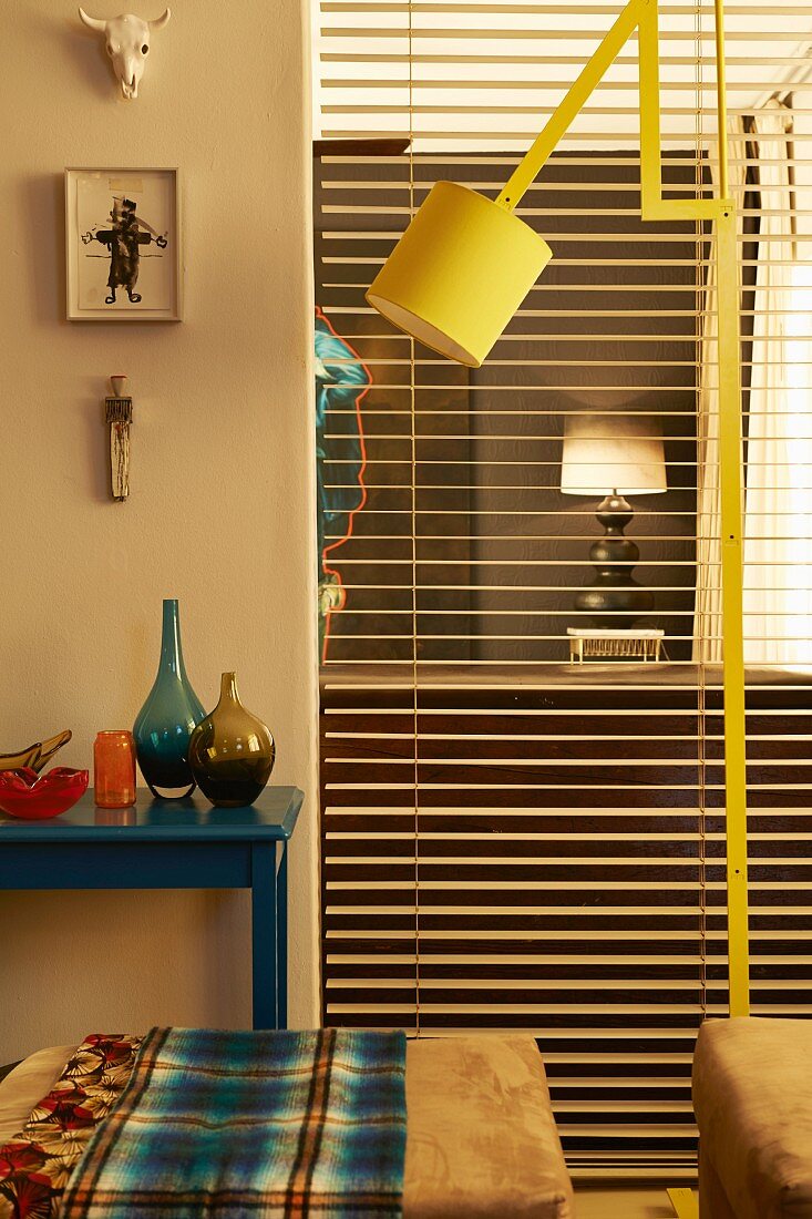 Yellow, adjustable standard lamp between ottomans and full-length, open louver blind with view into adjacent room