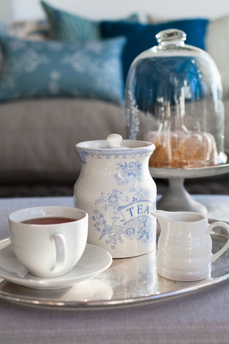 Teetasse, Milchkännchen und Teedose auf Silbertablett, im Hintergrund Kuchen unter Glashaube