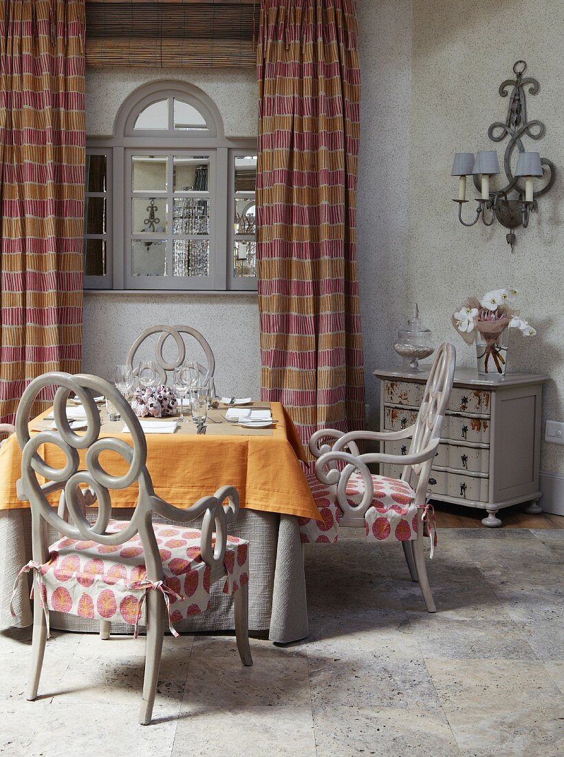 Gedeckter Tisch mit oranger Tischdecke und grau lackierte Stühle mit rosettenartigem Muster auf Rückenlehne, im Hintergrund Fenster mit gestreiften, bodenlangen Vorhängen
