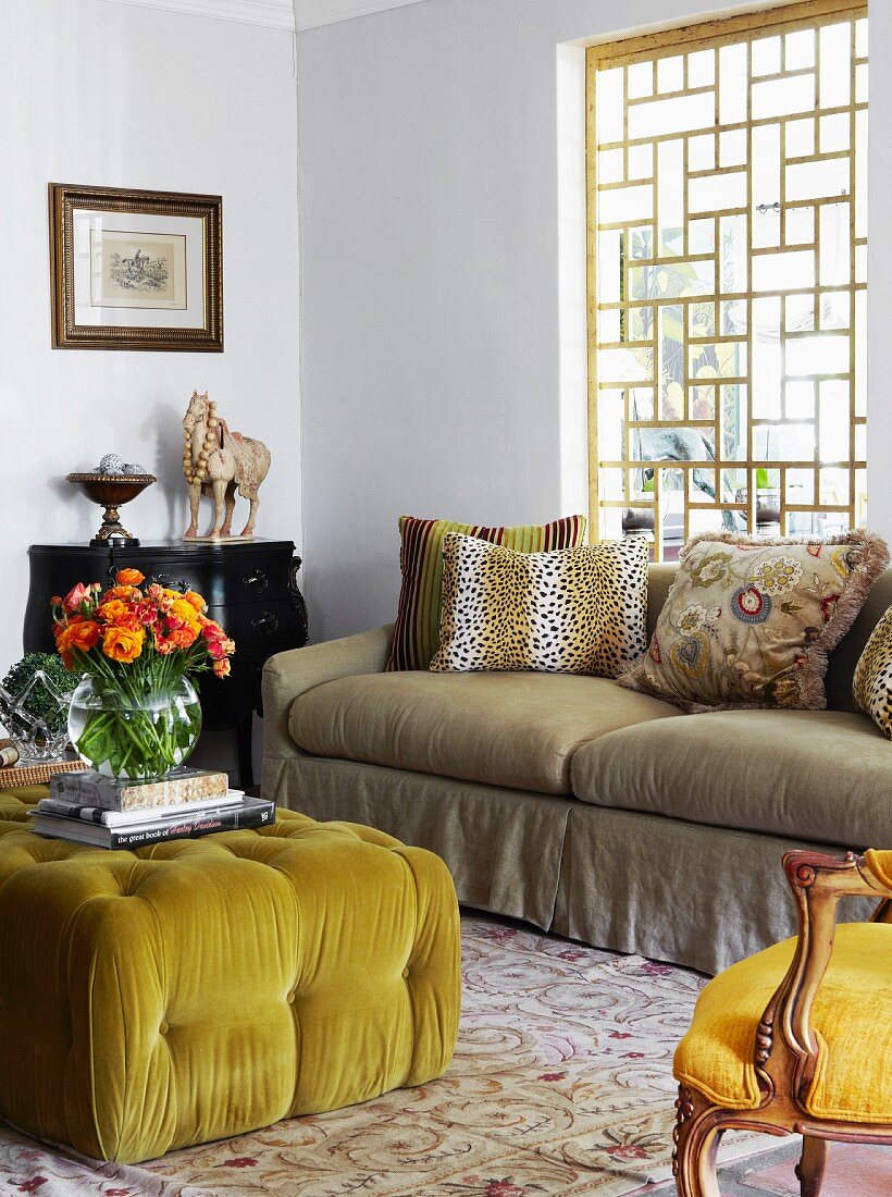 Gepolsterter Couchtisch mit gelbgrünem Bezug und bunter Blumenstrauss in Vase gegenüber Sofa am Fenster mit gitterartigem Holzgestell