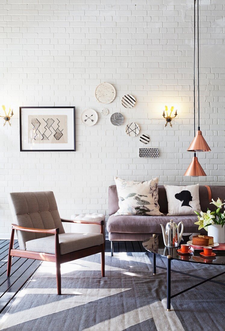Fifties armchair and grey sofa around coffee table below pendant lamps with copper-coloured lampshades in loft-style interior with decorative wall plates on whitewashed brick wall