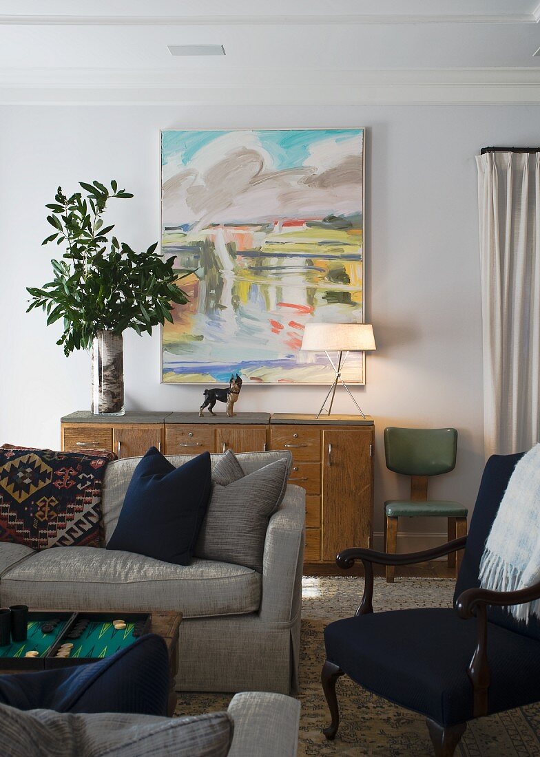Graue Couch vor Sideboard, darauf Vase mit Blätterzweigen und Tischleuchte, vor modernem Landschaftsbild
