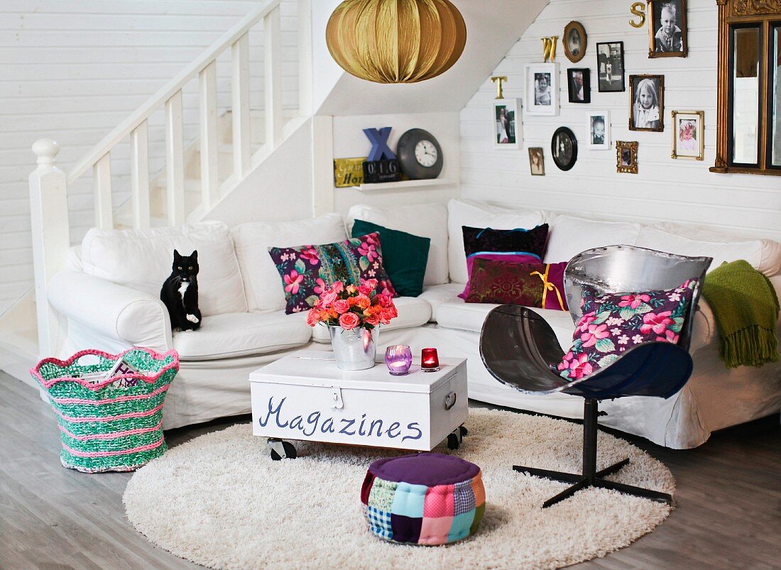 Colourful scatter cushions on white corner sofa and magazine box on castors used as coffee table; gallery of photos on wall and country-house staircase in background