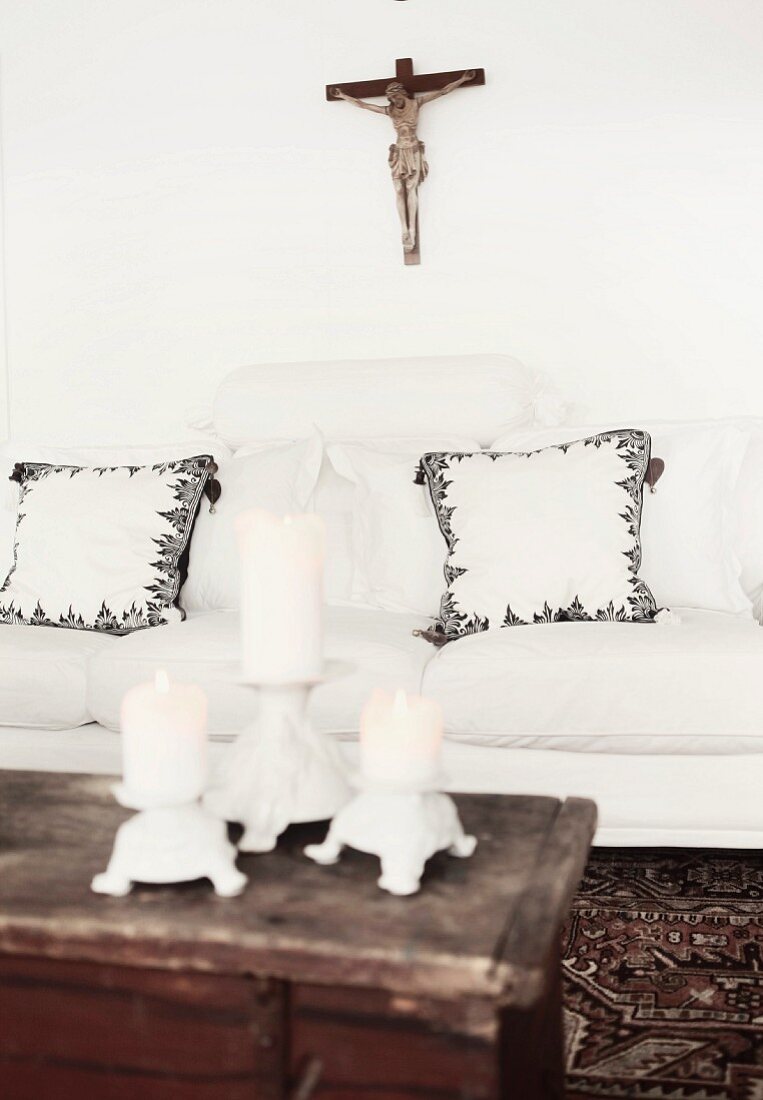 View across three candlesticks on wooden trunk to white sofa with scatter cushions