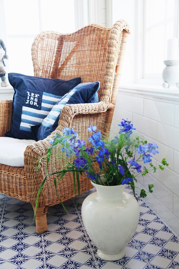 Blue cushion on wicker chair and vase of delphiniums on blue and white, patterned, tiled floor