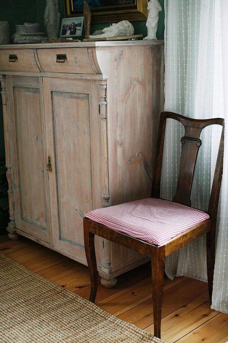 Antique wooden chair with seat upholstered in gingham next to pale wooden dresser