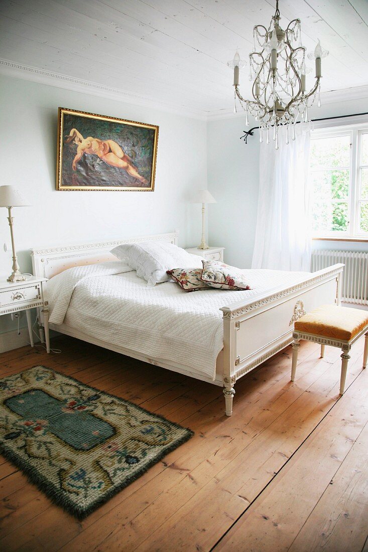 Simple bedroom with Regency-style double bed and bedside table painted white below chandelier wit glass pendants