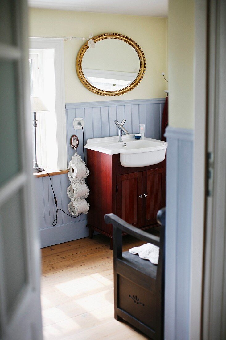 Blick durch offene Tür ins Bad, Waschtisch mit Unterschrank aus Mahagoniholz, darüber ovaler Spiegel mit Goldrahmen an hellgelb getönter Wand
