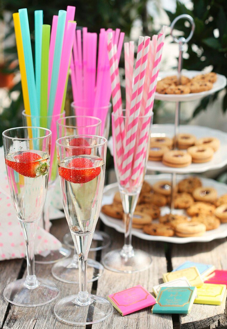 Champagne with strawberries in flutes on garden table; colourful straws and cake stand of biscuits in background