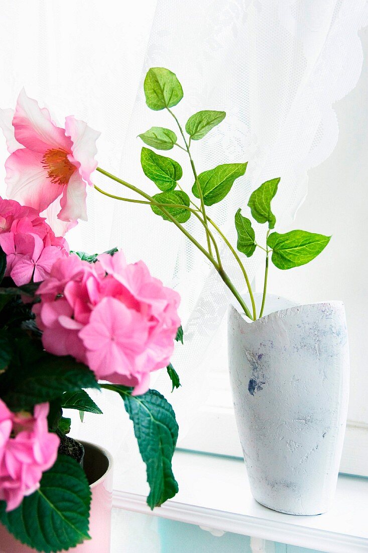 Weiß lasierte Vase mit pinkfarbener Blume, seitlich Zimmerpflanze mit rosa Blüten