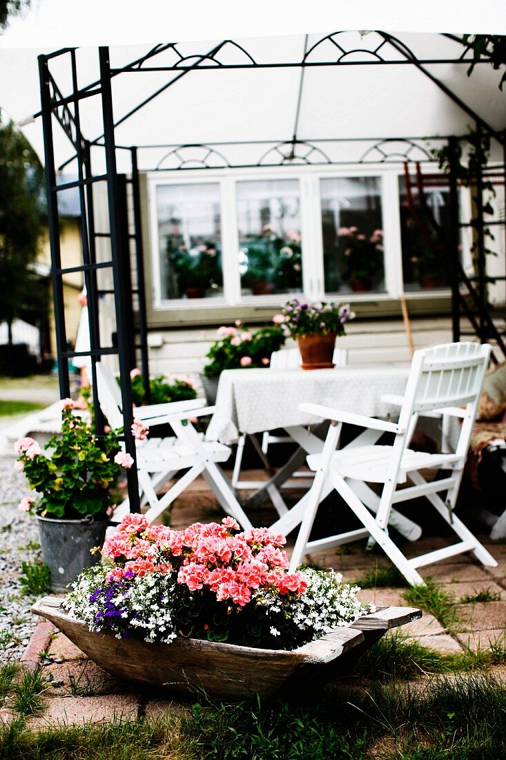 Terrassenplatz mit weiss lackierten Holzstühlen um Tisch, unter Rankgerüst als Pavillon, davor Holzschale mit Blumen auf Boden