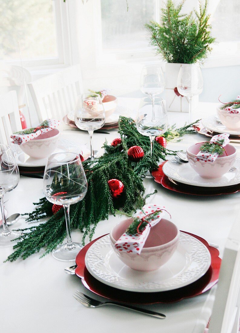 Für ein festliches Mittagessen gedeckter Tisch mit mittig drapiertem Koniferenzweig und roten Weihnachtsbaumkugeln